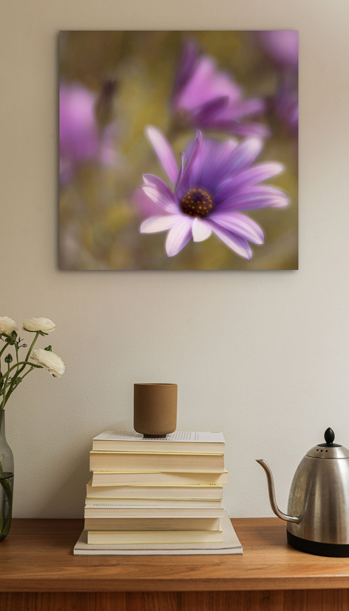 Picture hanging on a wall. There is also a desk with a stack of books. The picture is a fine art photograph of a purple daisy blowing the in the wind. The photograph is by Cameron Dreaux. 