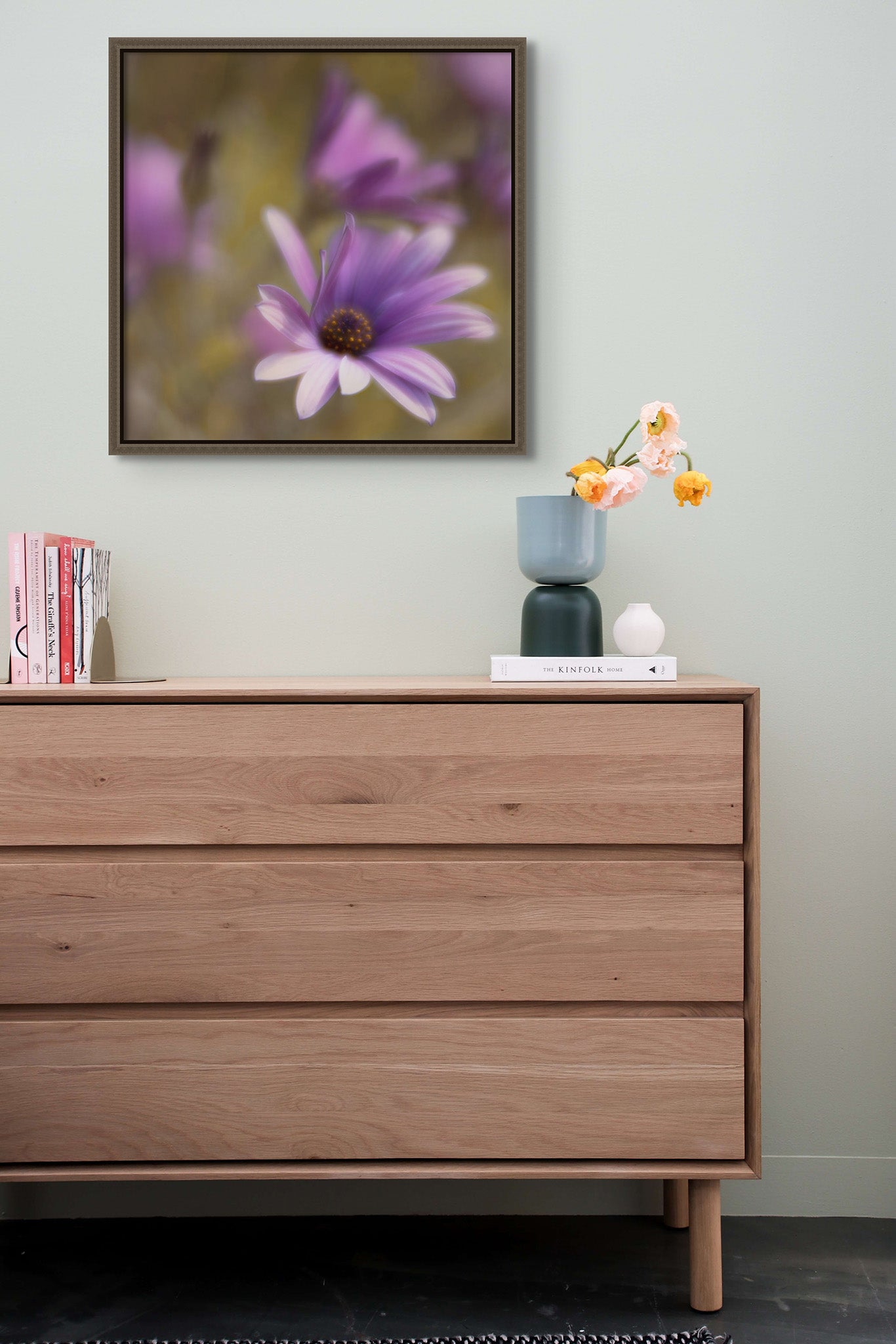 Picture hanging on a wall. There is also a dresser with flowers in a vase. The picture is a fine art photograph of a purple daisy blowing the in the wind. The photograph is by Cameron Dreaux. 