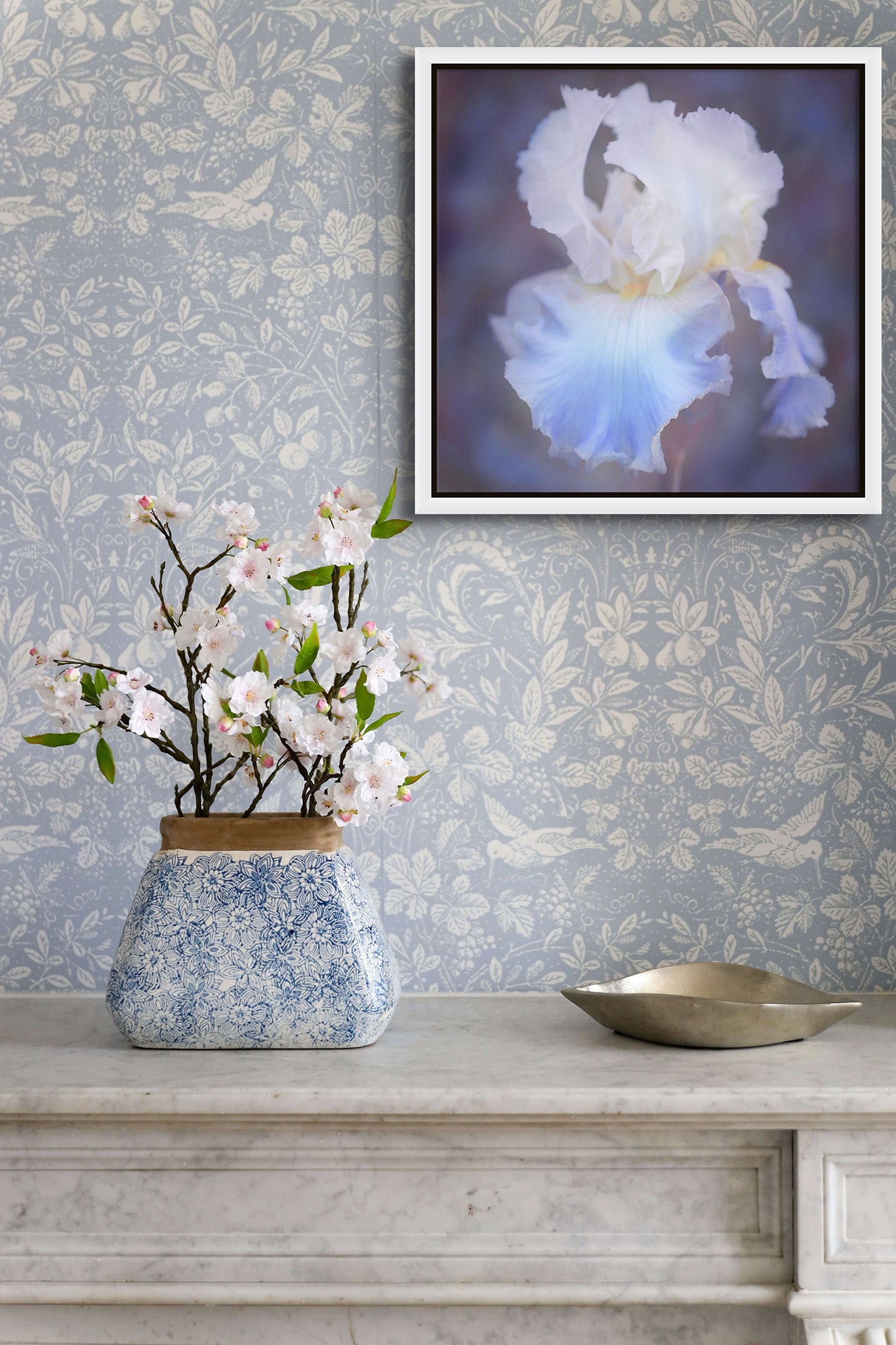 Picture hanging on the wall of a room. There is a counter with a bouquet of flowers. There is ornate wallpaper on the all. The picture is a blue and white dahlia photograph by Cameron Dreaux. 