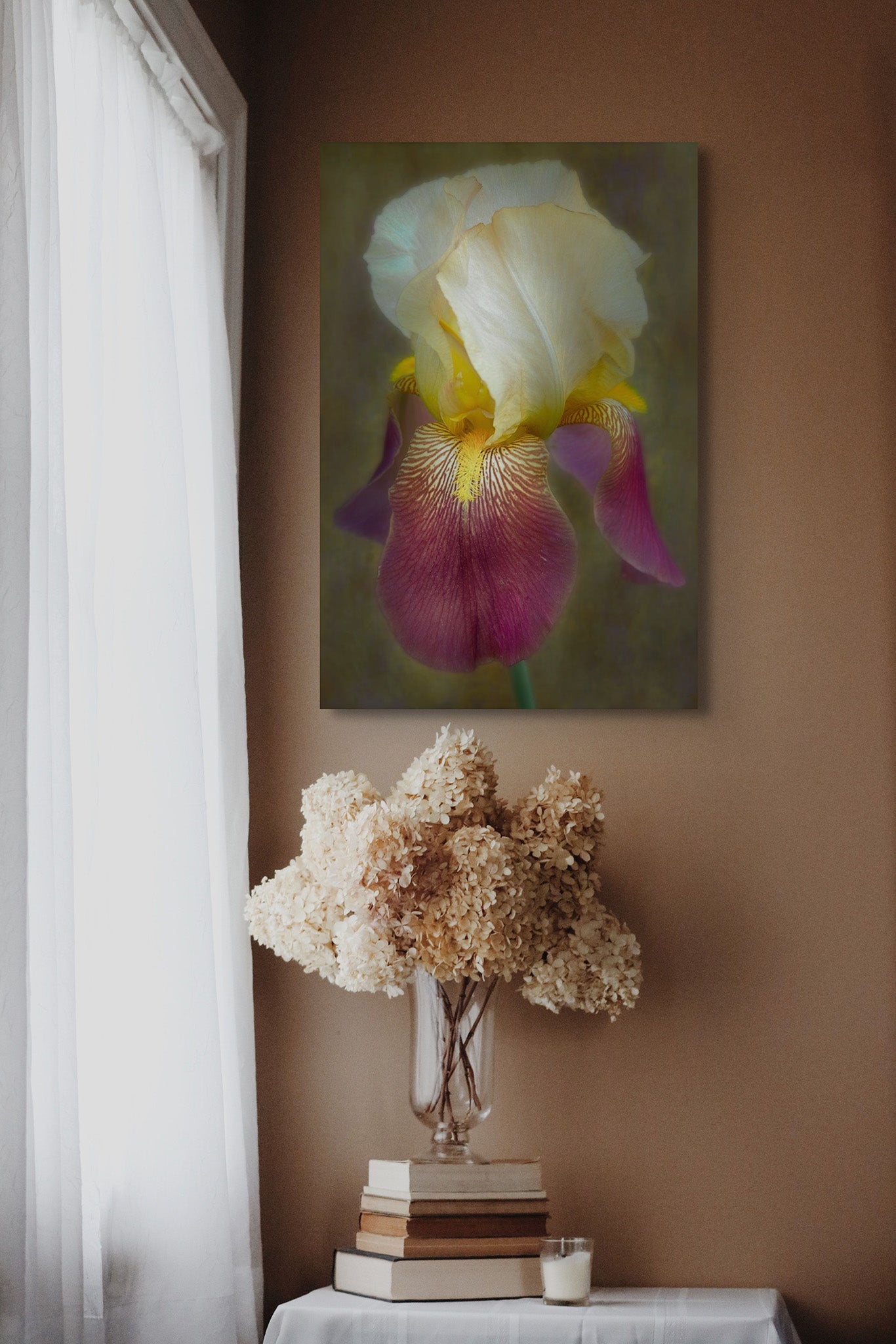 A picture hanging on the wall of a room by a window. There is a bouquet of flowers below the picture in a clear vase. The picture on the wall is a photograph of an Iris by Cameron Dreaux. 
