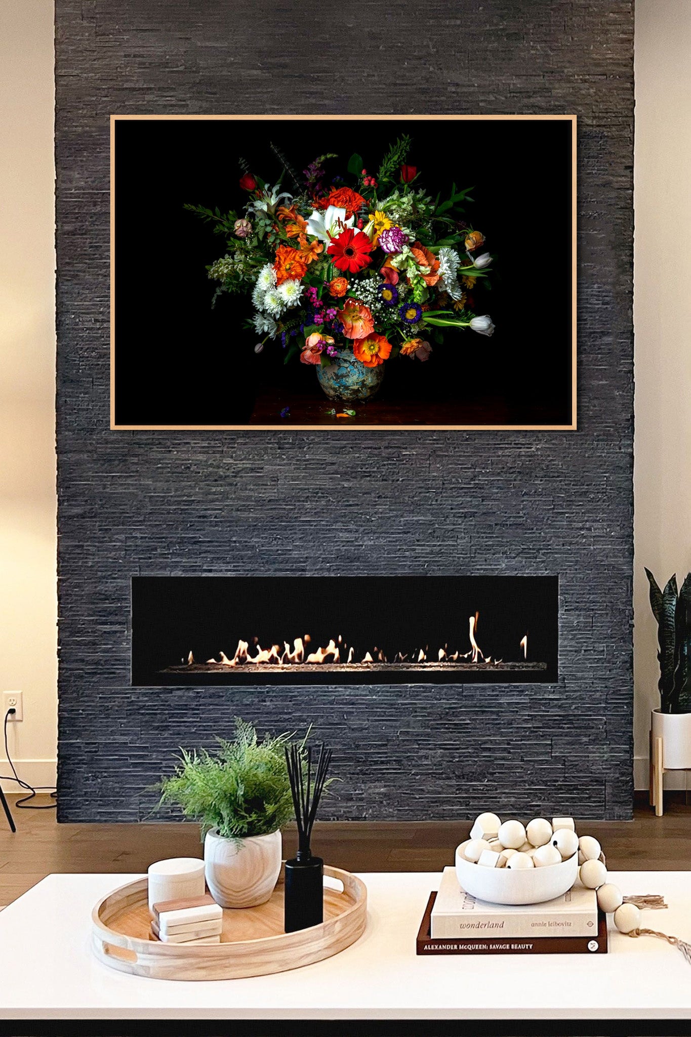A picture hanging on the wall of a living room. There is a table and fireplace with a fire burning.. The picture is a metal print from Dreaux Fine Art of "A Generous Gift." "Generous Gift" is a photograph of a bouquet of various types of flowers on a black background.