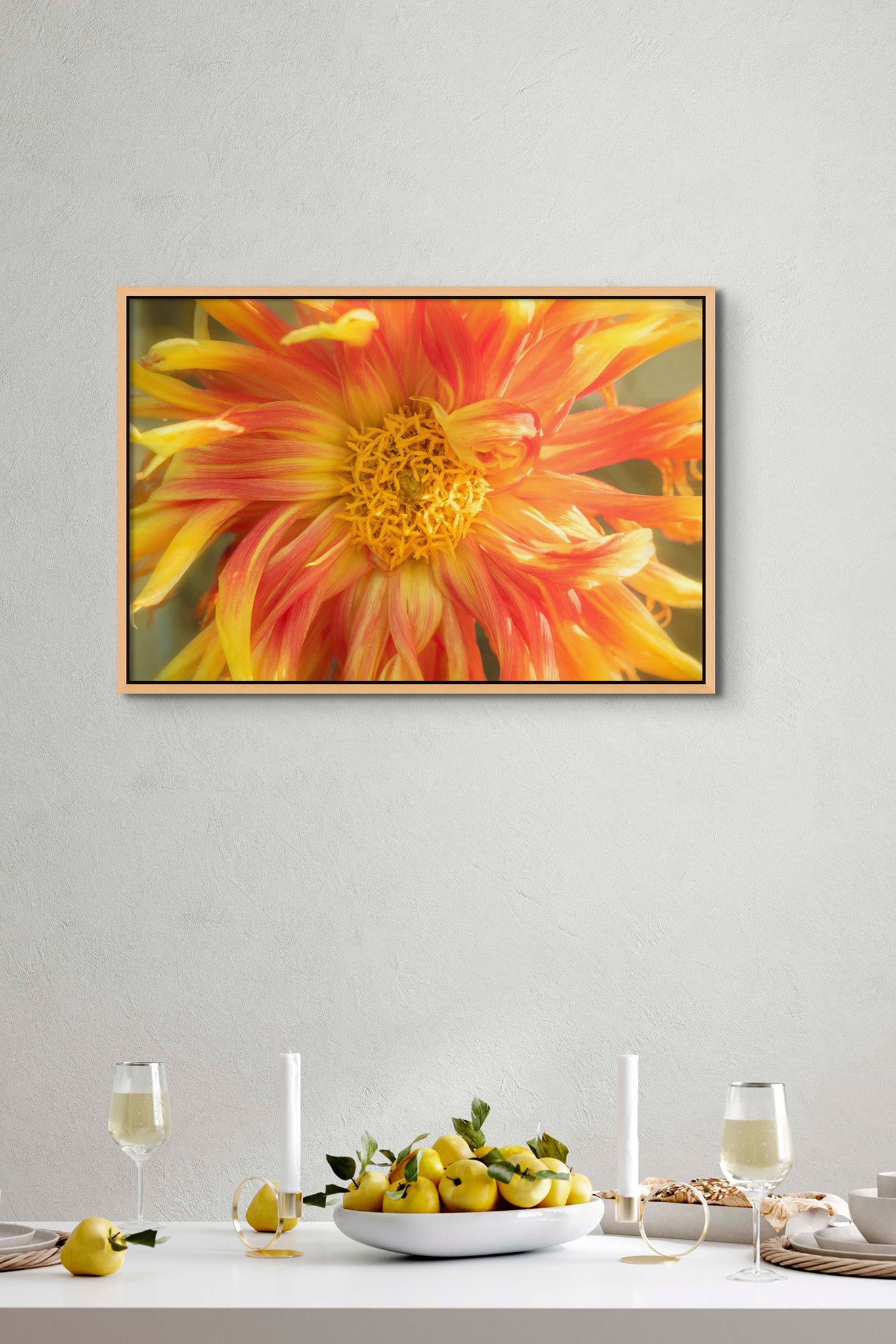 Picture hanging on wall of a dining room with a set table. On the table is glasses of wine and a bowl of lemons. The picture is a photograph of a flowing yellow dahlia flower by Cameron Dreaux. 