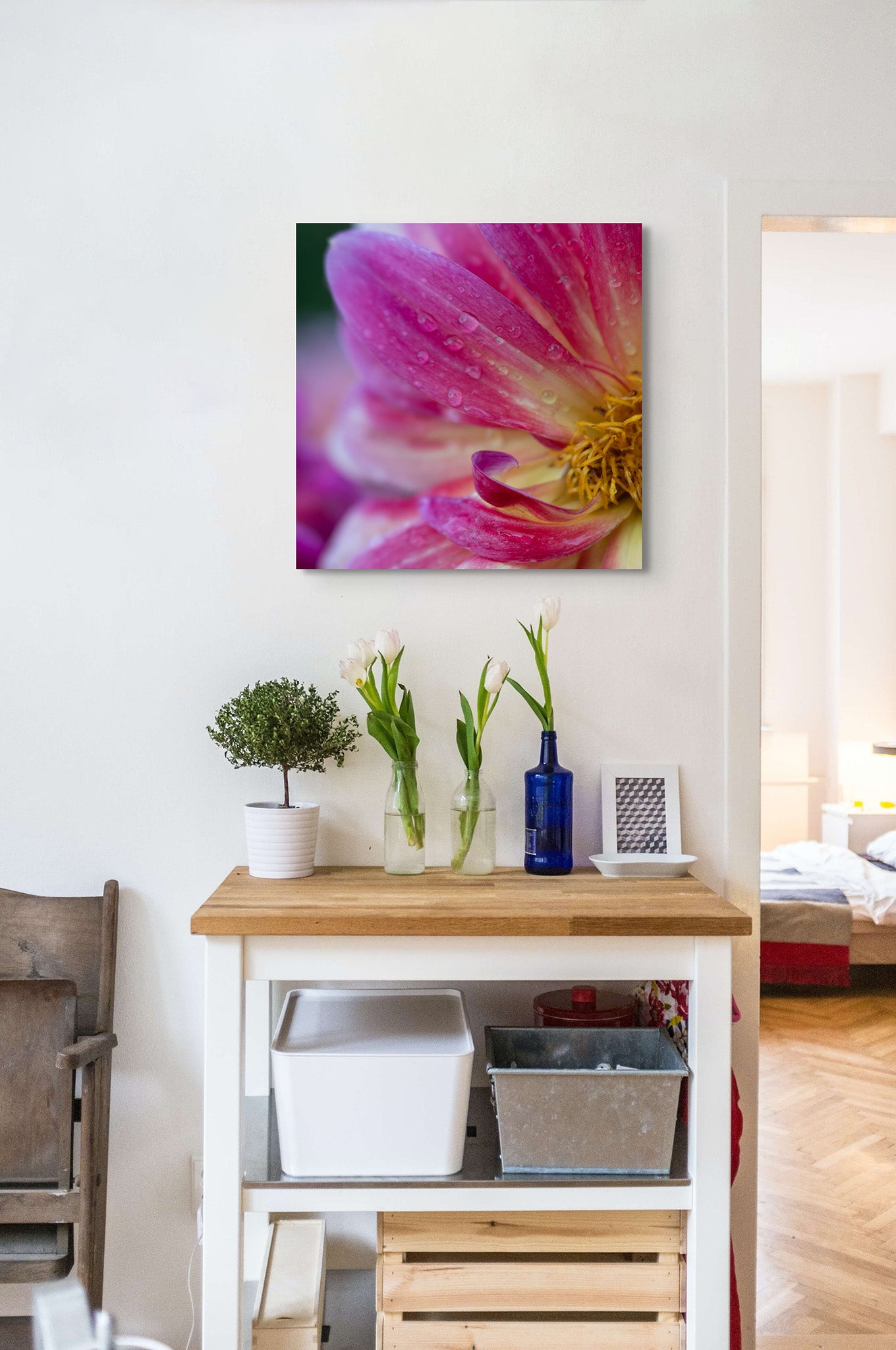 Picture hanging on the wall of a room. It looks like a kitchen. There is a wooden cart with flowers in vases. The picture on the wall is a fine art macro photograph of a pink Dahlia flower by Cameron Dreaux. 