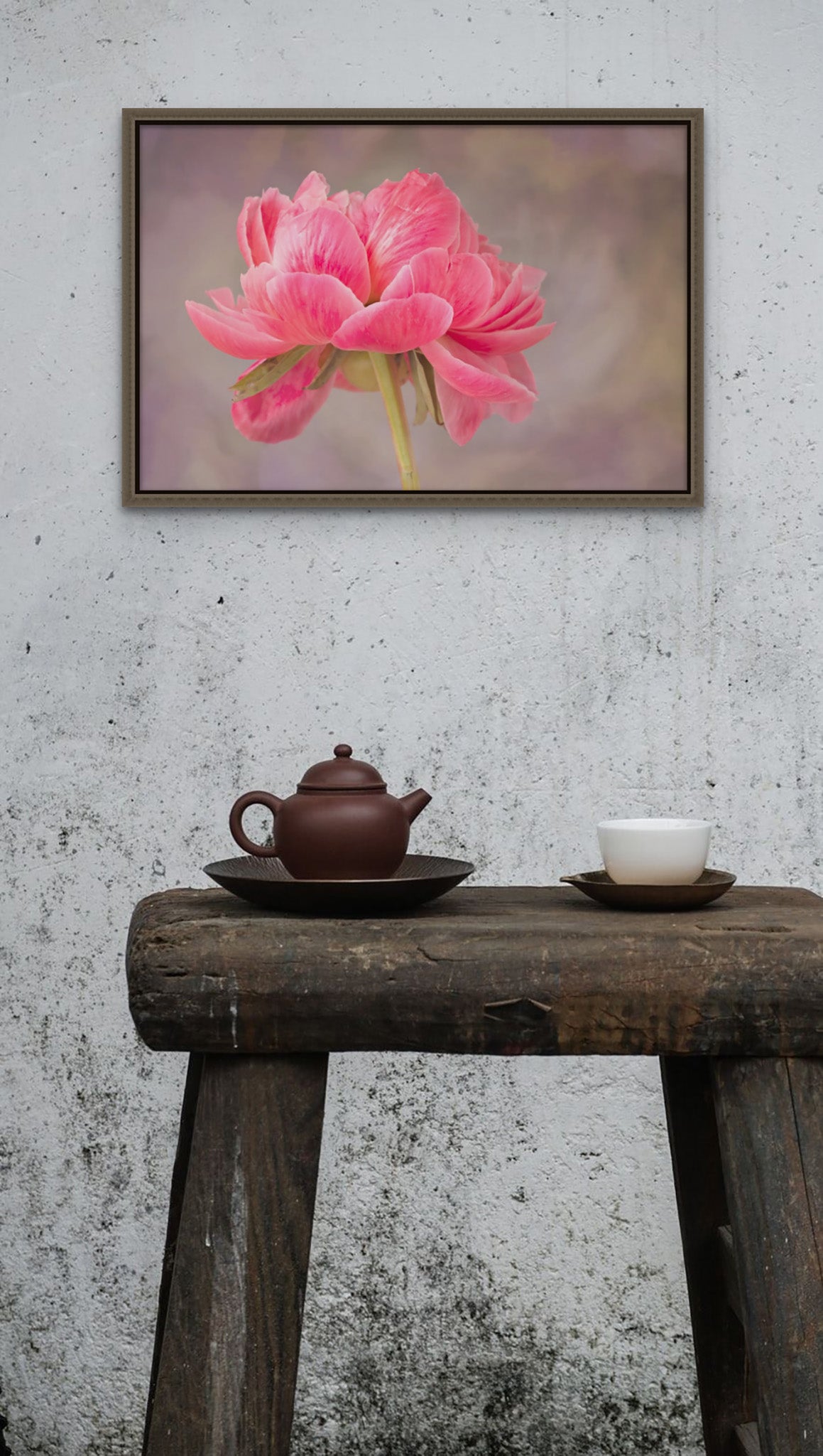 Picture hanging on the wall of a room with a rustic table. The photograph is in a float frame. The photography is a fine art Metal print of pink Peony with the petals blowing in the wind. The photograph is by Cameron Dreaux. 