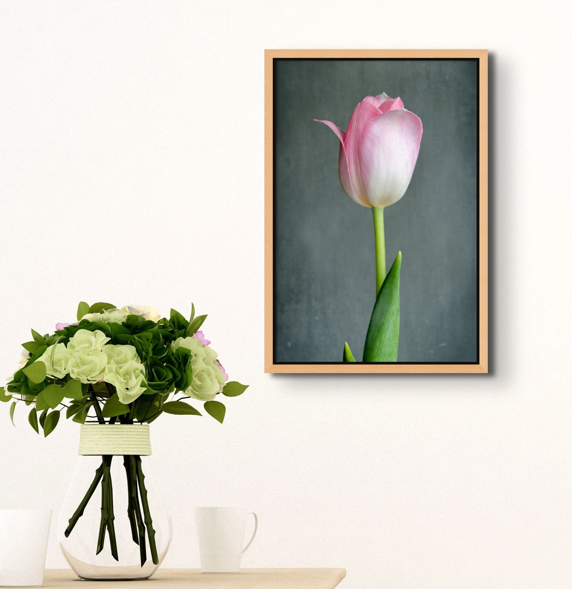 Picture with a floating frame hanging on the wall of a room. There is a table below the picture. On the table is a bouquet of flowers in a vase.The picture is a fine Art Photograph of a singular pink tulip flower on a grey background by Cameron Dreaux. 