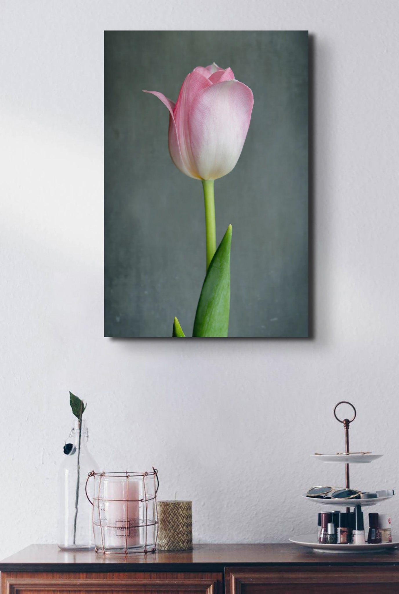 Picture hanging on the wall of a room. There is a table below the picture. The picture is a fine Art Photograph of a singular pink tulip flower on a grey background by Cameron Dreaux. 