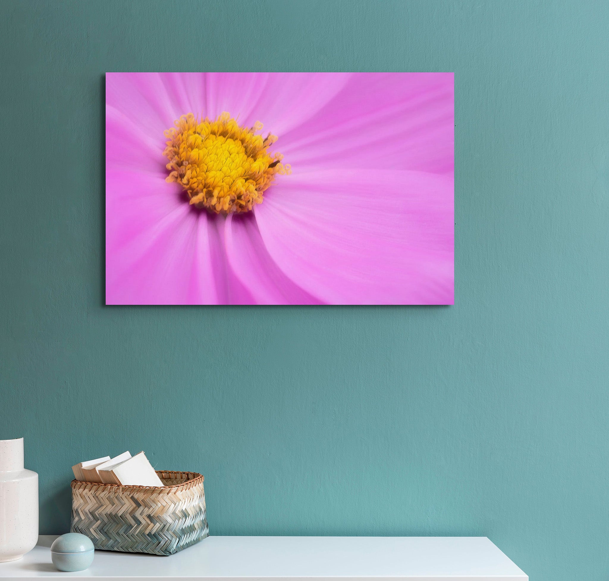 Picture hanging on a green wall with a countertop underneath. The countertop has a basket on it and some other miscellaneous items. The picture is a macro photograph of purple cosmopolitan flower by Cameron Dreaux. 